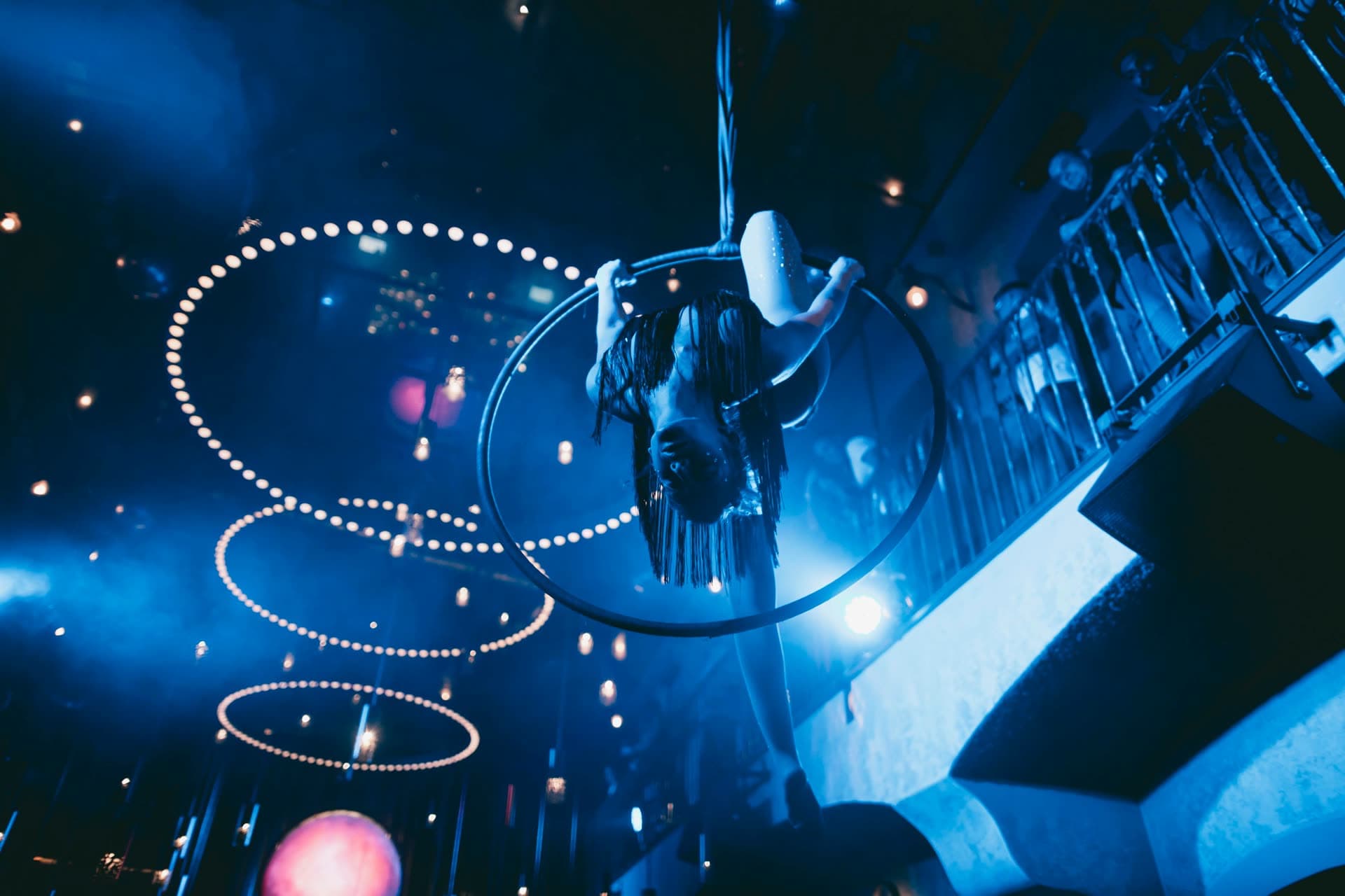 Aerial hoop – inverze na vzdušném kruhu v modře nasvíceném klubu, světelné spirály v pozadí
